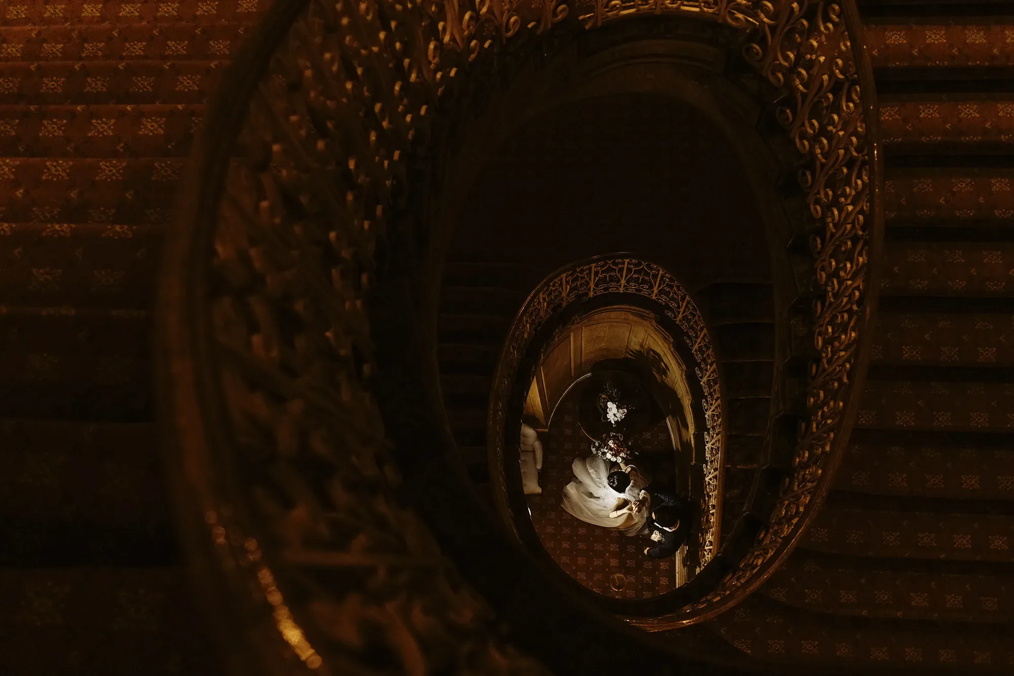 An ornate, spiral staircase at the Fairmont Olympic Hotel, viewed from above and illuminated by soft lighting, with a person captured in motion at the bottom standing next to a statue. captured by Seattle Wedding Photographers Jenn Tai & Co