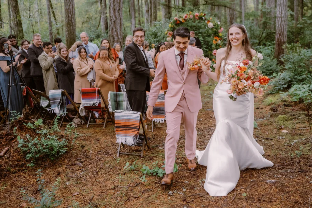 A bride and groom walk down a forest aisle post-ceremony, smiling, with guests watching from either side. the bride holds a large bouquet, and the groom is in a pink suit. captured by Seattle Wedding Photographers Jenn Tai & Co