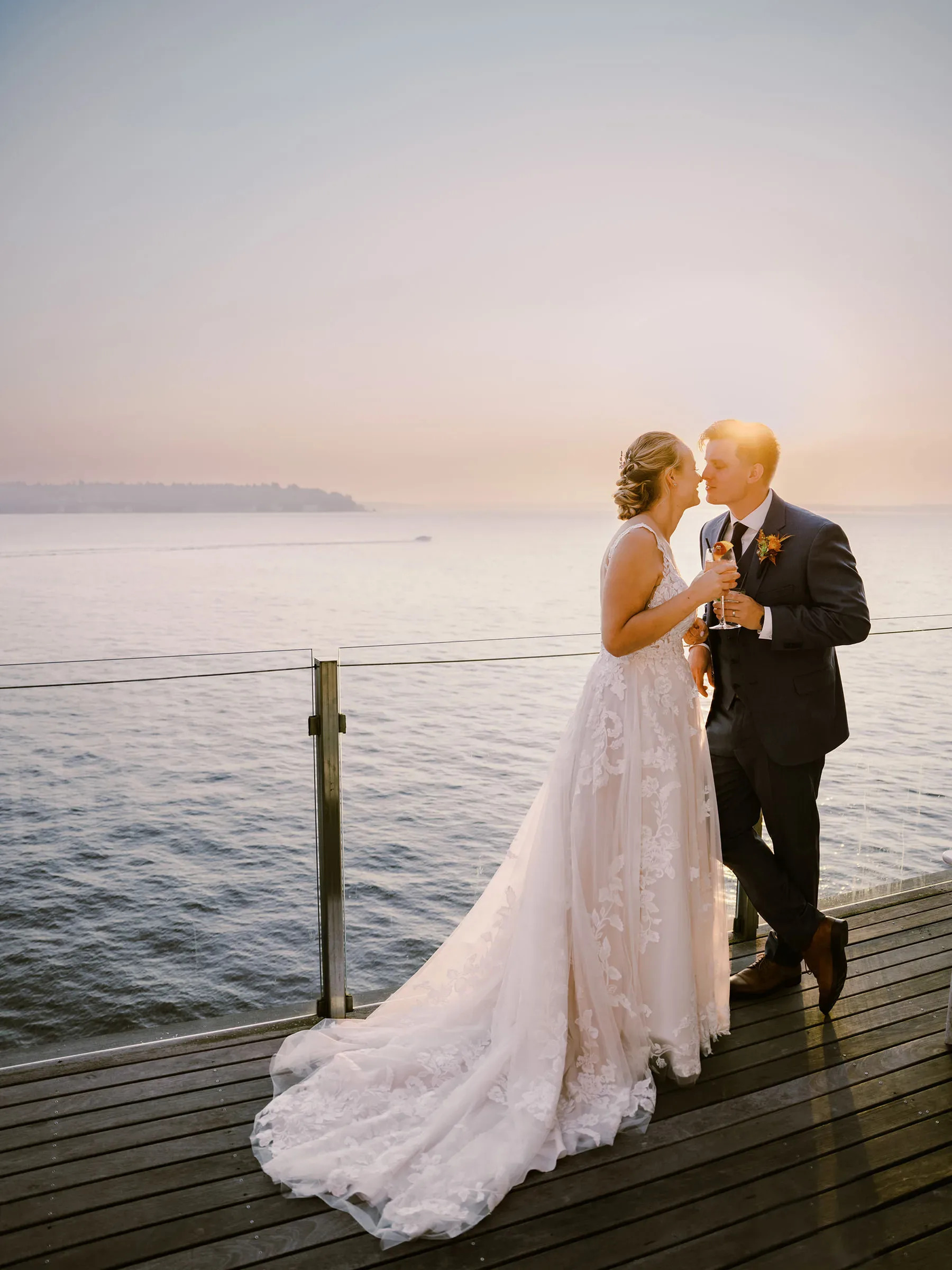 A bride and groom share a romantic moment on a dock at sunset, kissing gently with glasses of champagne in hand, surrounded by a serene ocean backdrop. captured by Seattle Wedding Photographers Jenn Tai & Co