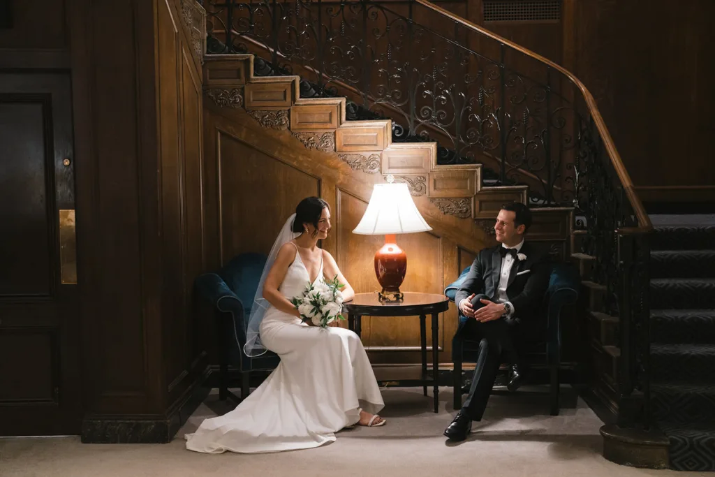 A bride in a white dress and a groom in a black tuxedo sit at a small table under a lamp, beside an ornate wooden staircase at the Fairmont Olympic Hotel, engaged in conversation captured by Seattle Wedding Photographers Jenn Tai & Co