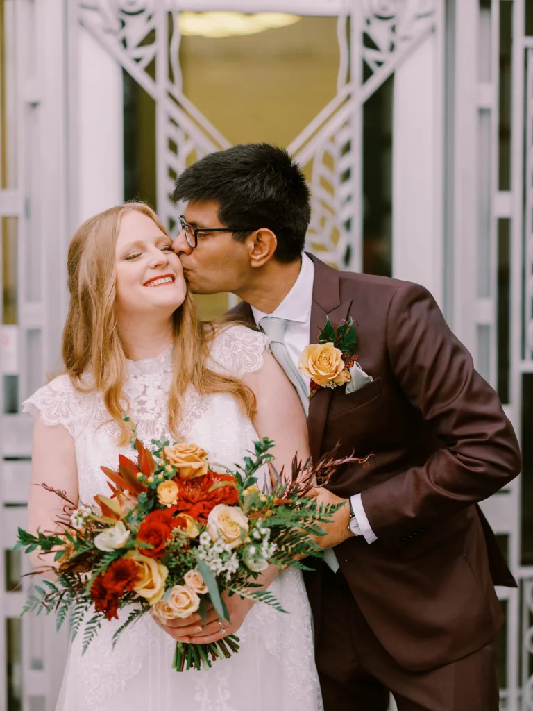 A groom in a brown suit is kissing the cheek of a bride holding a bouquet of red and orange flowers; they are standing in front of the Seattle Asian Art Museum.