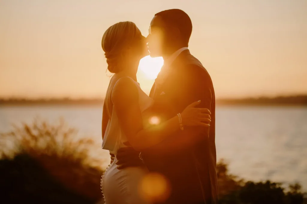 Bride and groom sharing a kiss at sunset by a lakeside, with warm backlighting and lens flare enhancing the romantic atmosphere. captured by Seattle Wedding Photographers Jenn Tai & Co