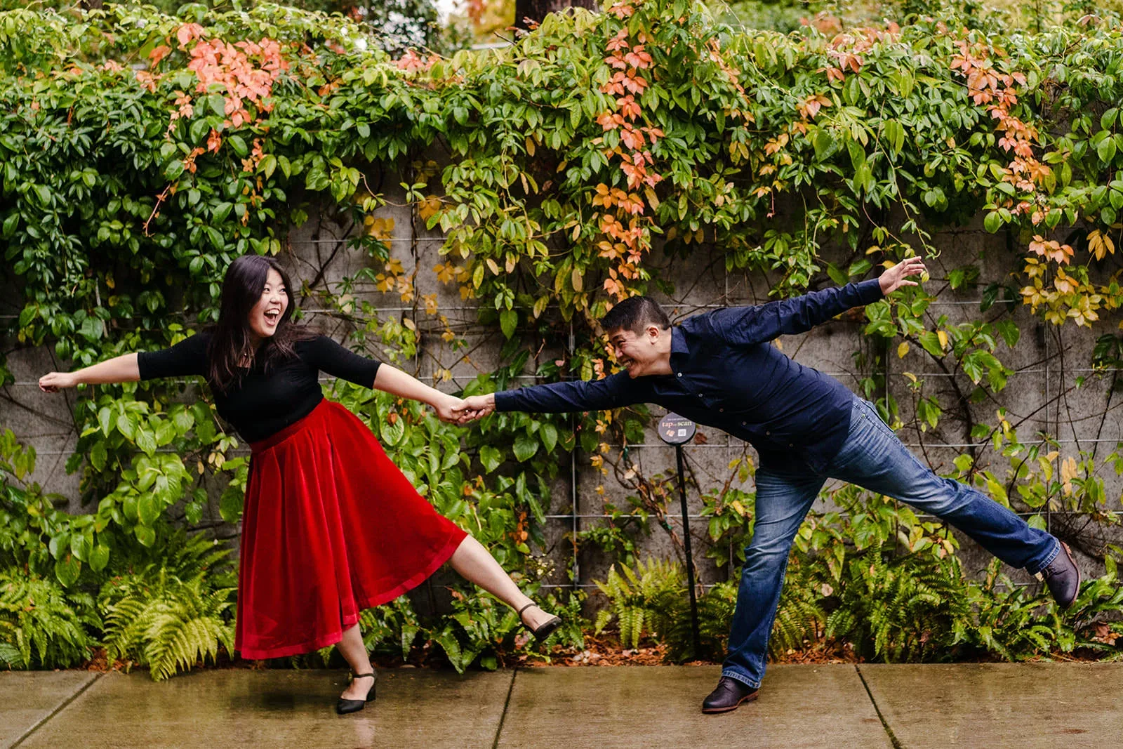 A man and woman hold hands while posing playfully on a wet pavement. The woman, in a red skirt and black top, leans back with a joyful expression. The man, in a dark blue shirt and jeans, extends his arms and one leg. A lush green and orange vine-covered wall is behind them. captured by Seattle Wedding Photographers Jenn Tai & Co