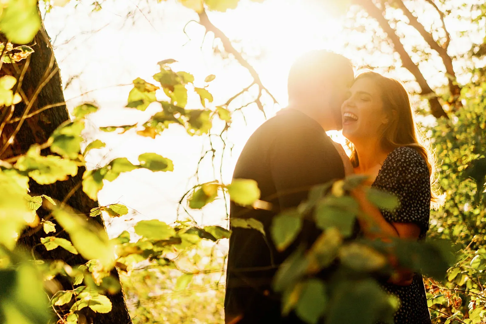 A couple stands in a sunlit forest, framed by green leaves. The man, in a black shirt, faces with his back partially to the camera and is whispering or kissing the woman's forehead. The woman, in a dark patterned dress, smiles brightly. The sunlight creates a warm backlight effect. captured by Seattle Wedding Photographers Jenn Tai & Co