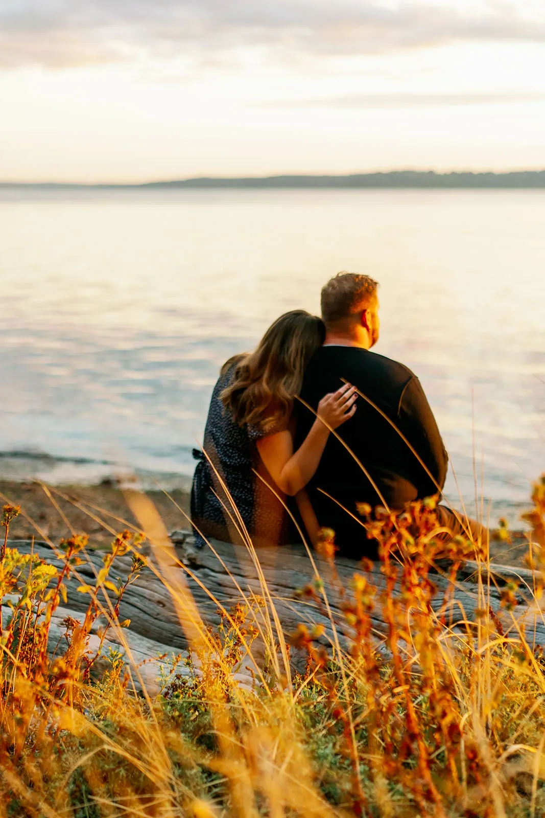 A couple sits on a log by the water's edge, with the woman resting her head on the man's shoulder. The scene is bathed in the warm, golden light of sunset. Tall grasses and wildflowers frame the foreground, adding to the serene atmosphere. captured by Seattle Wedding Photographers Jenn Tai & Co