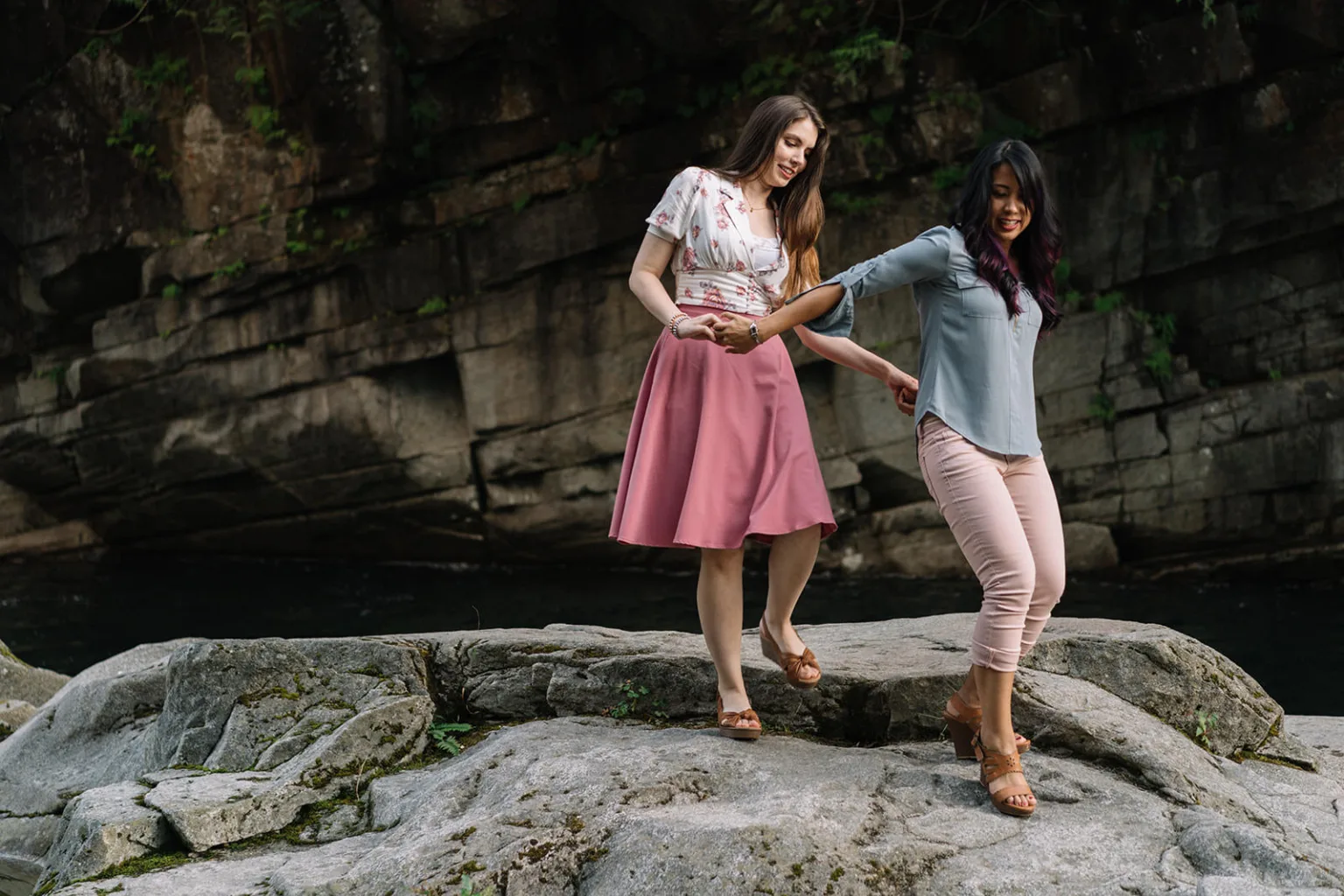 Two women walking hand in hand on a rocky terrain. One wears a floral blouse and pink skirt, while the other wears a blue top and pink pants. They navigate large stones with a background of a rocky wall. Both appear engaged and cautious. captured by Seattle Wedding Photographers Jenn Tai & Co